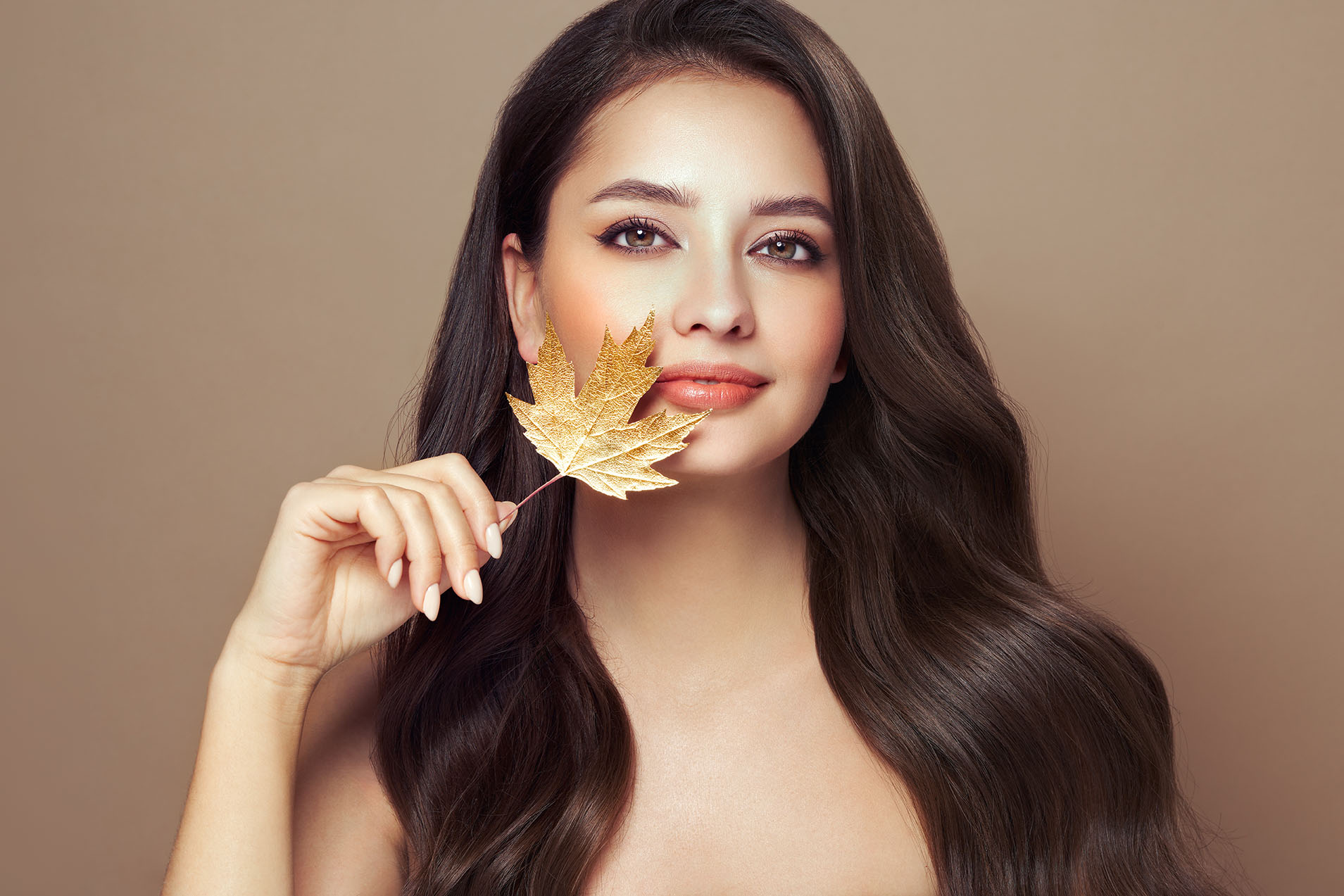 woman with long dark hair holding a gold maple leaf near her face against a warm brown background