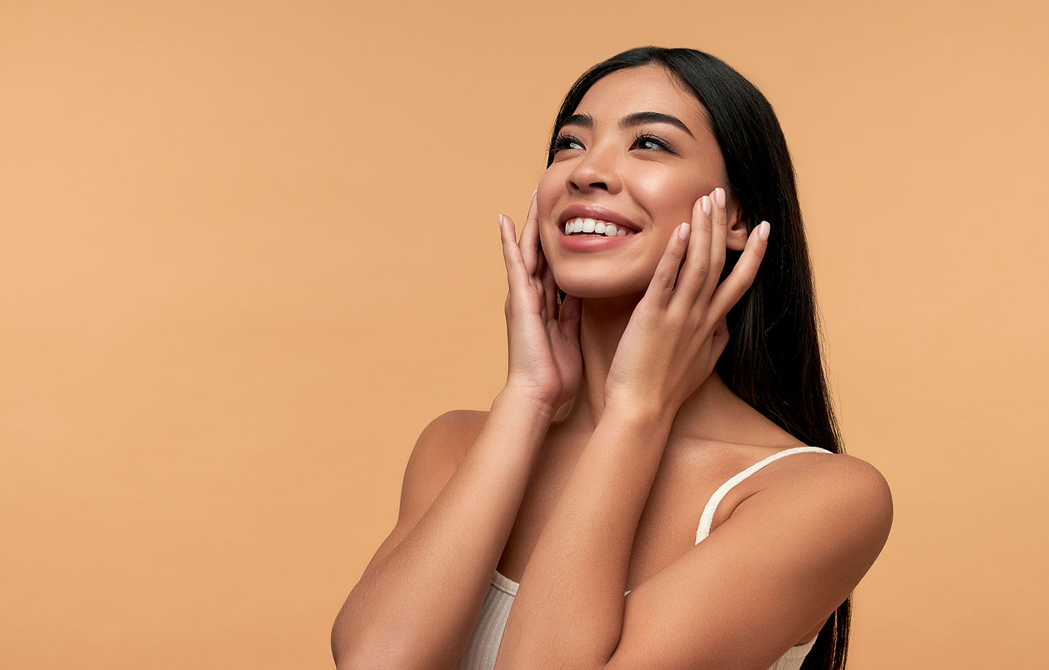 woman with long dark hair smiling and touching her cheeks with both hands against a beige background