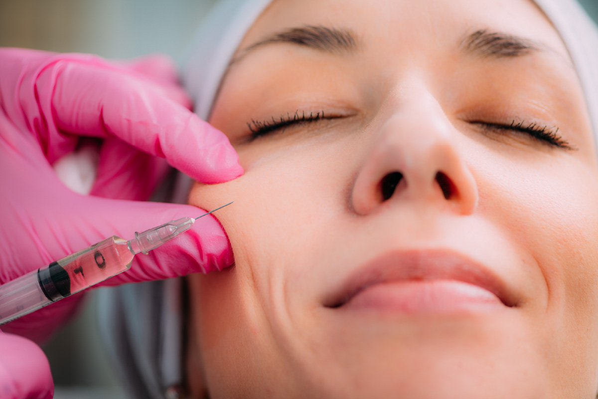 gloved hands holding a syringe to a woman’s cheek as she lies with eyes closed