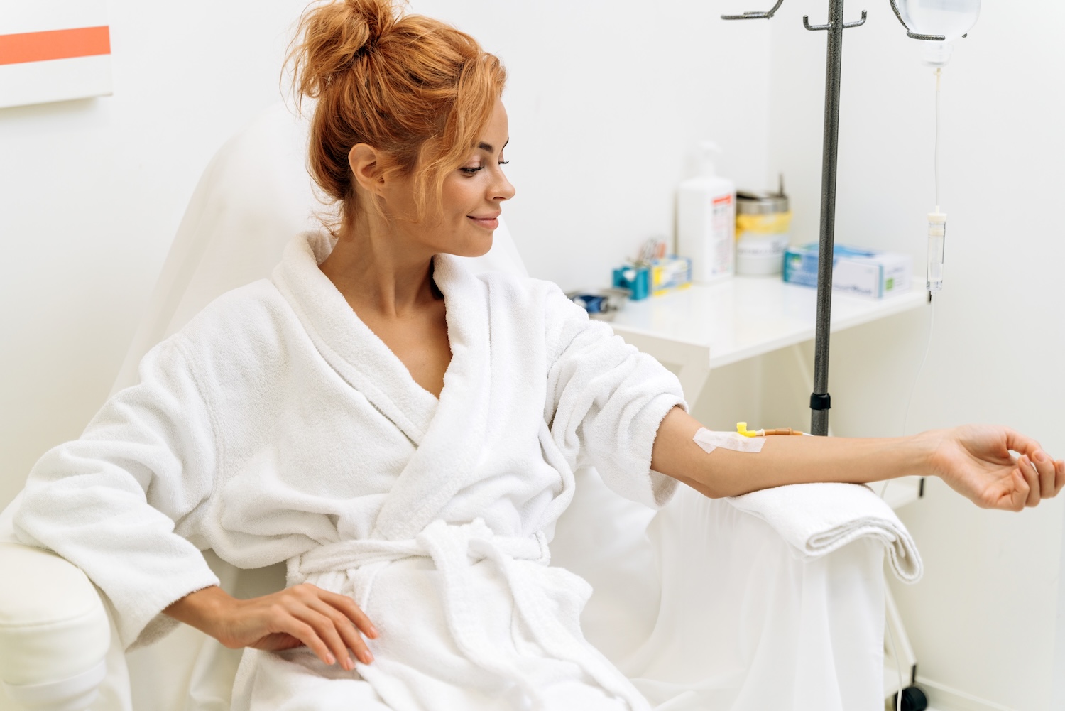 woman sitting in a chair wearing a white robe with an IV line taped to her arm and an IV bag hanging beside her