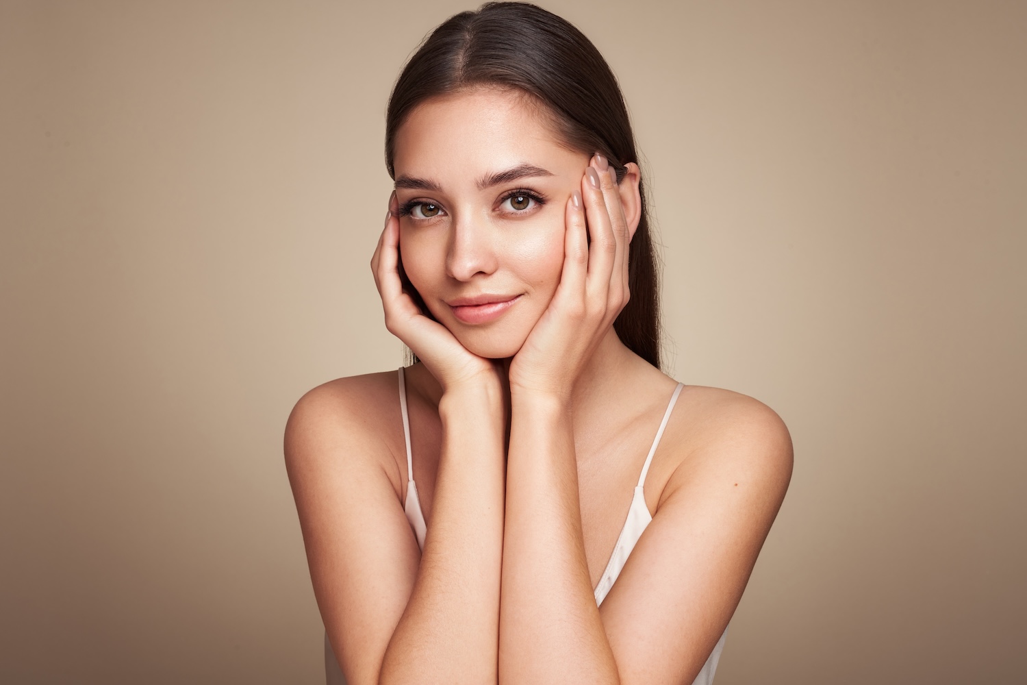 woman with long dark hair touching her face with both hands against a neutral background