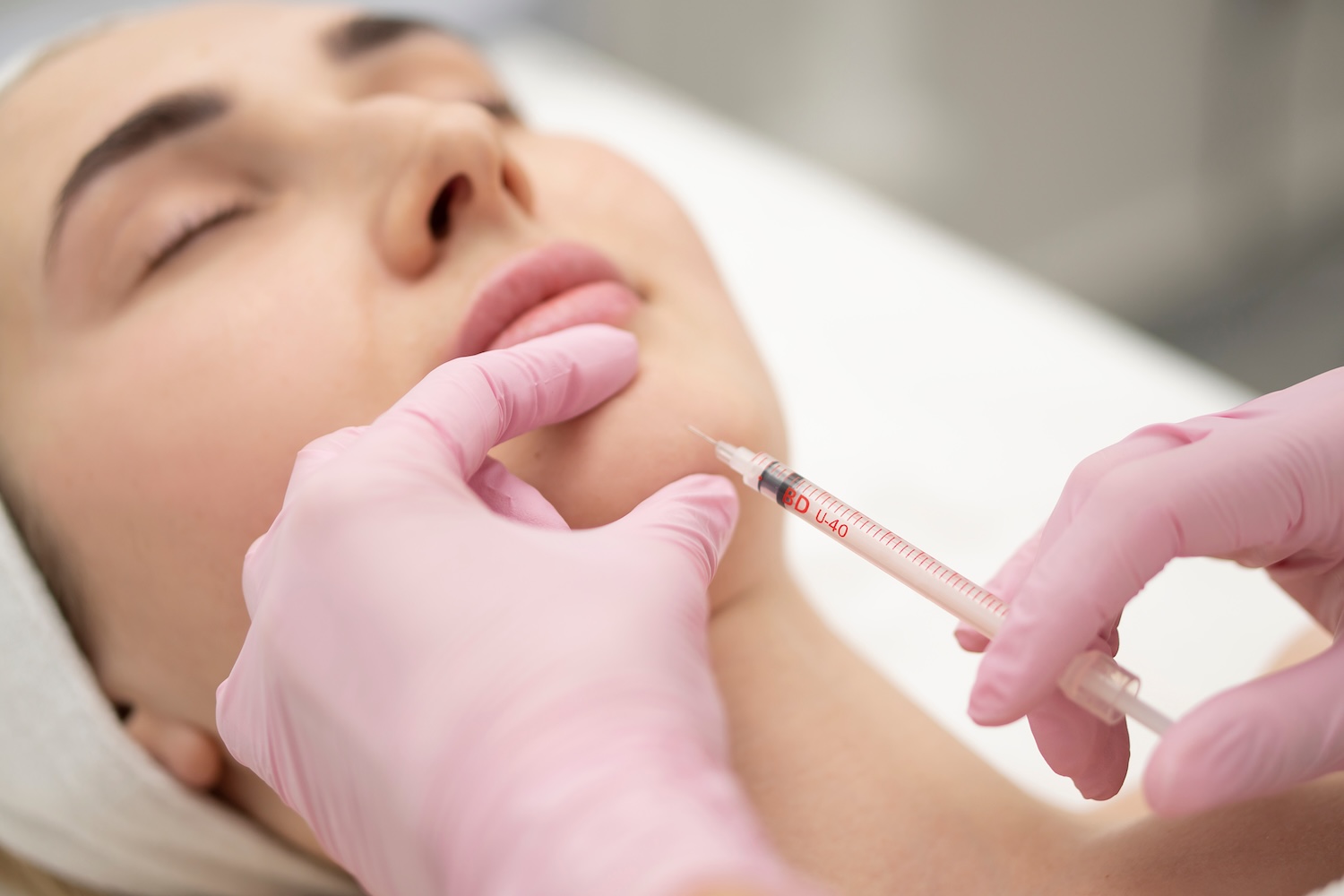 gloved hands holding a syringe near a woman’s chin as she lies with her eyes closed