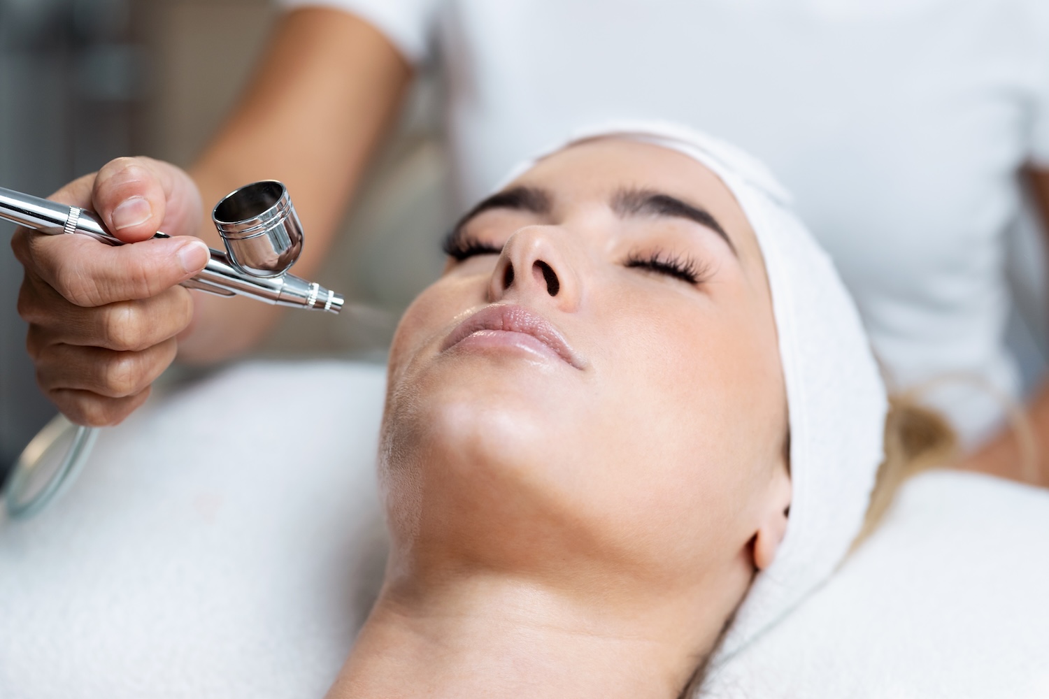 aesthetician performing an oxygen infusion facial using a handheld airbrush-style device on a client’s face