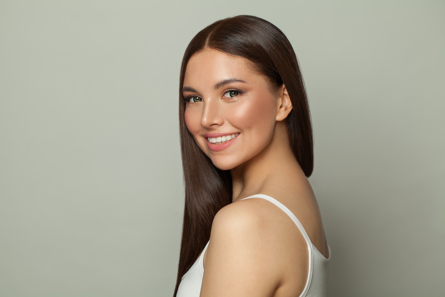woman with long straight hair smiling while looking over her shoulder against a plain background