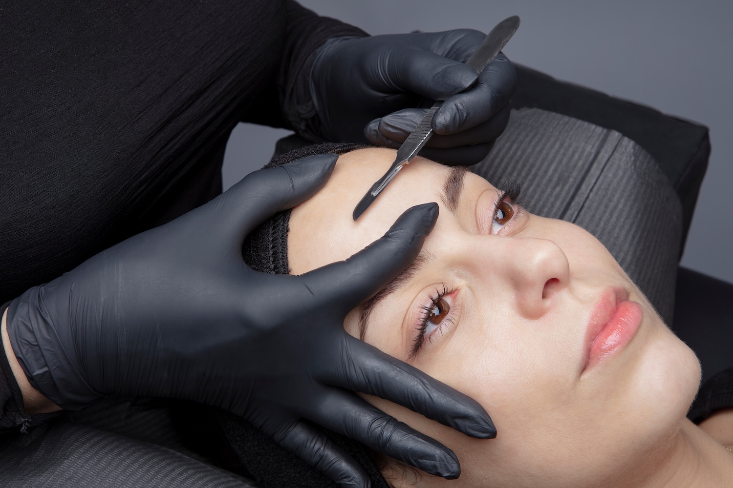 esthetician performing dermaplaning on a woman's face using a surgical scalpel for exfoliation and peach fuzz removal