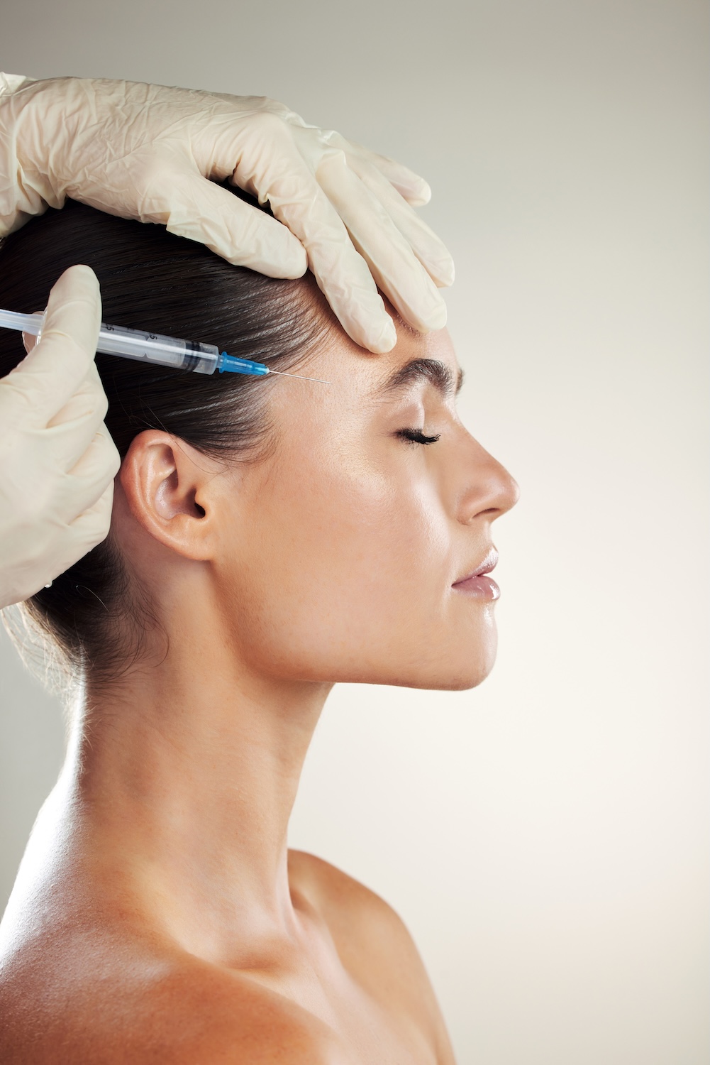 gloved hands holding a syringe near a woman’s temple as she faces sideways with eyes closed