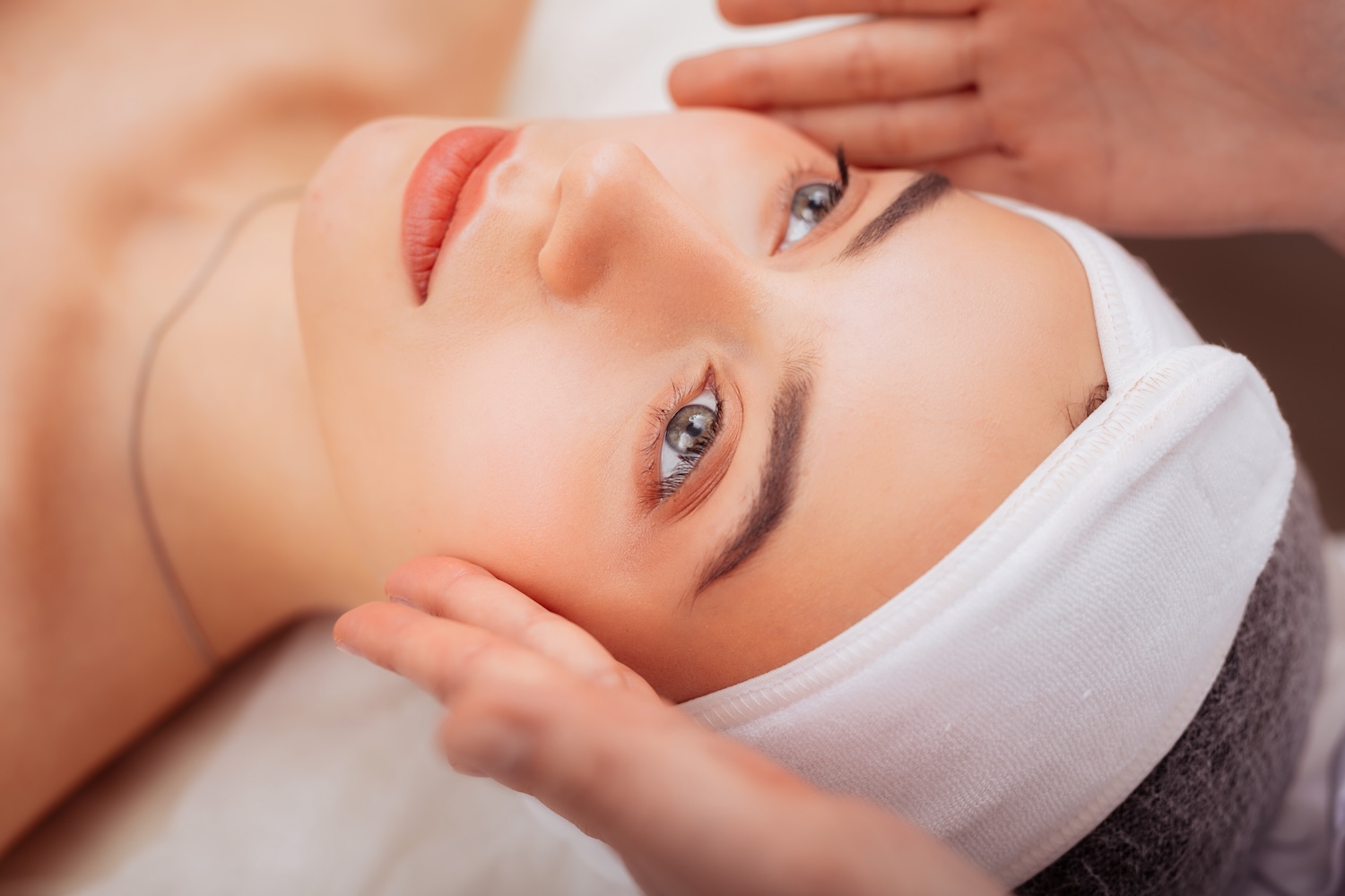 person lying with a white headband as hands hover near their temples during a facial treatment