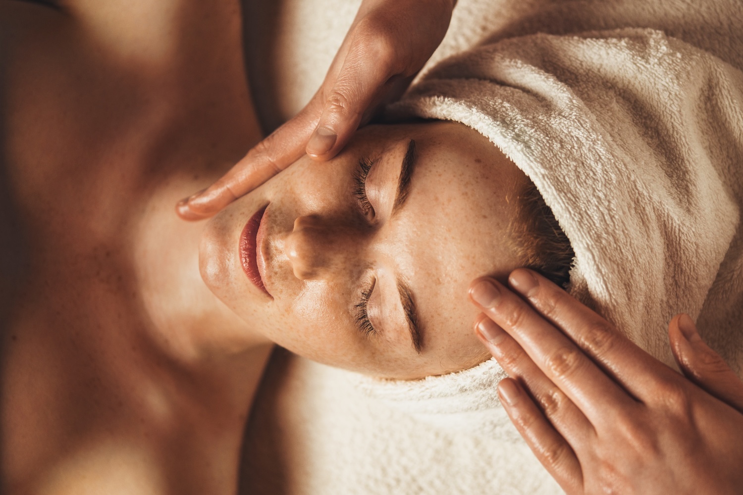 hands massaging the forehead of a person lying down with a towel wrapped around their head