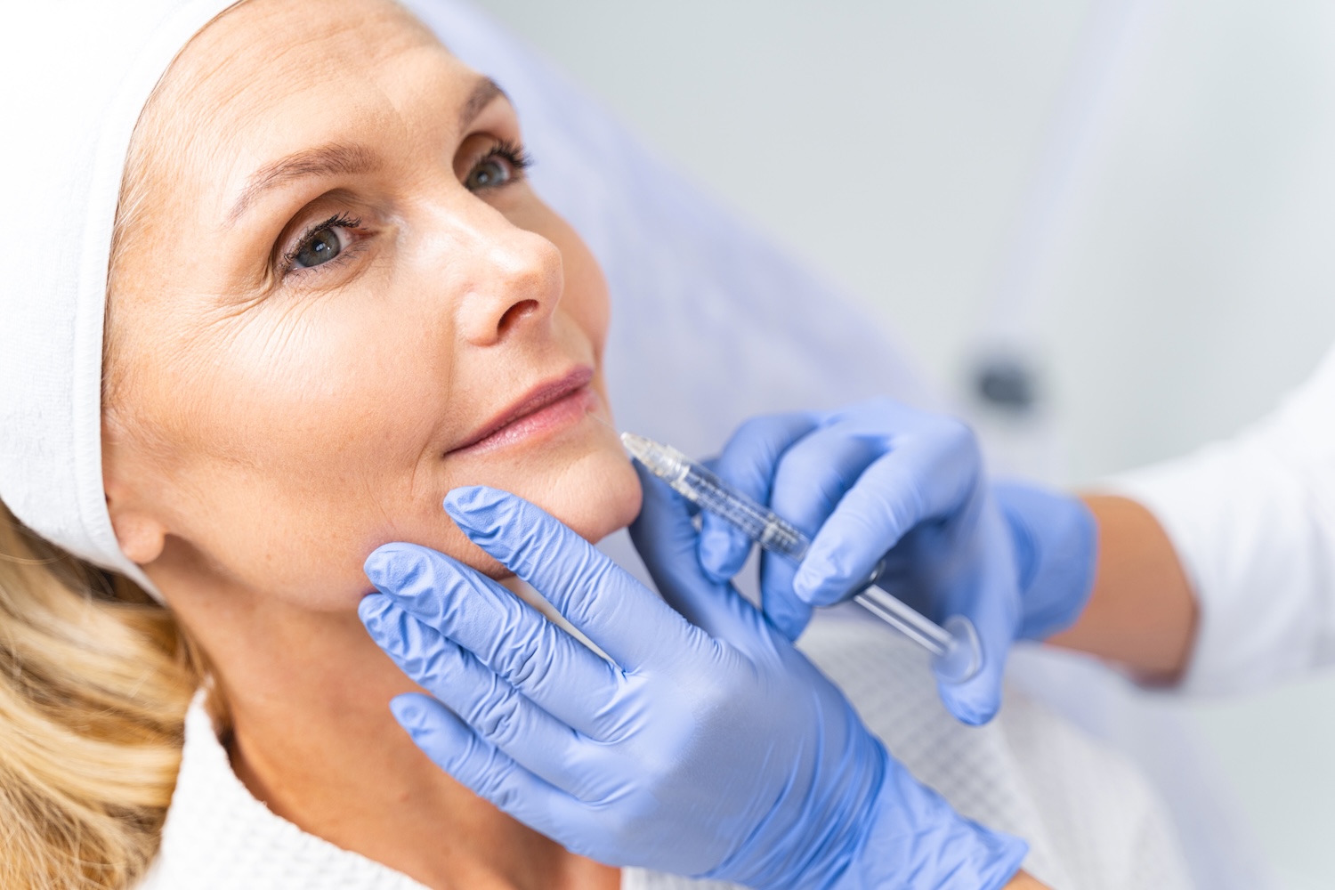 gloved hands holding a syringe at a woman’s lower cheek while she sits with a headband on
