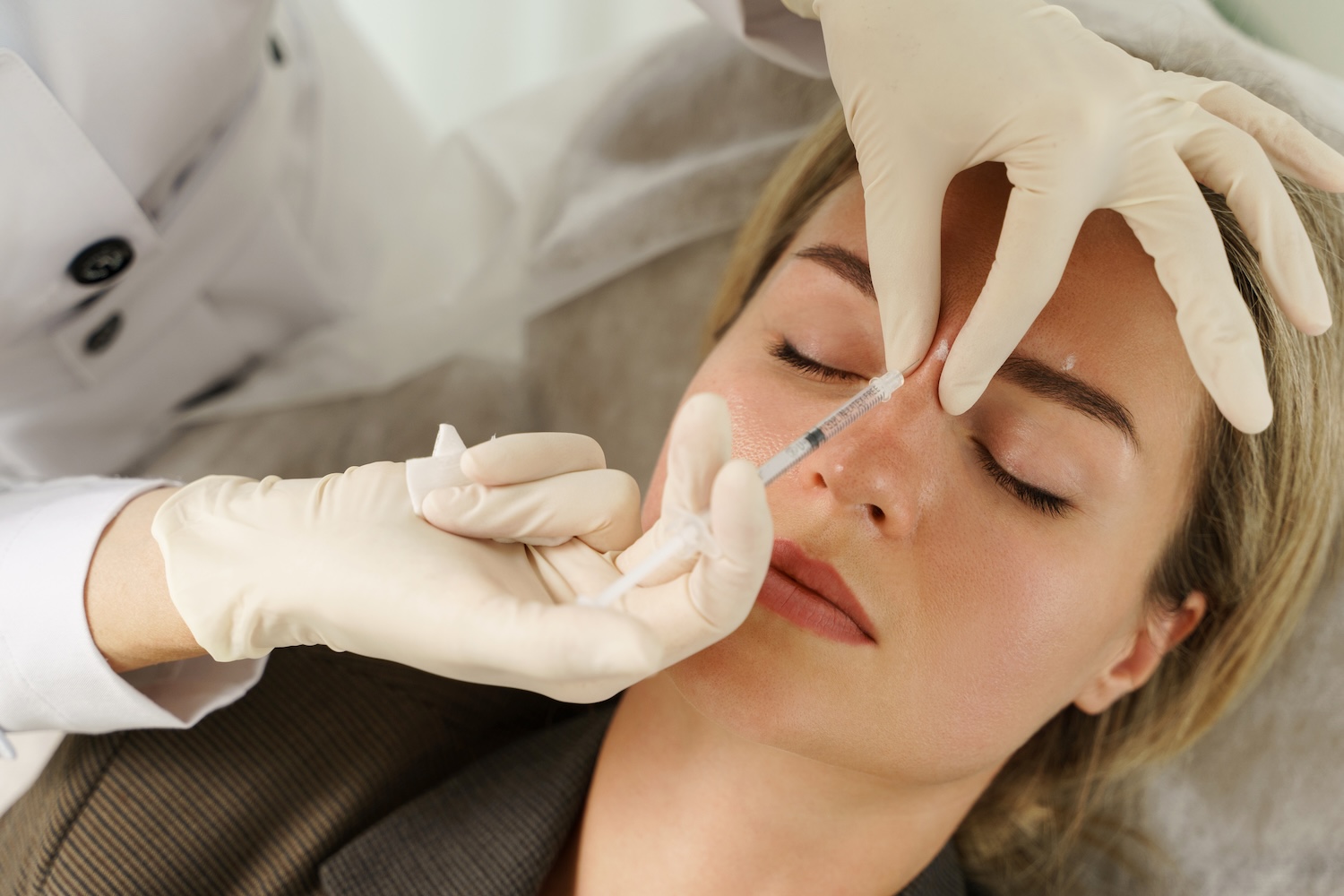Gloved hands holding a syringe at the bridge of a woman’s nose while she lies on a treatment bed with eyes closed.