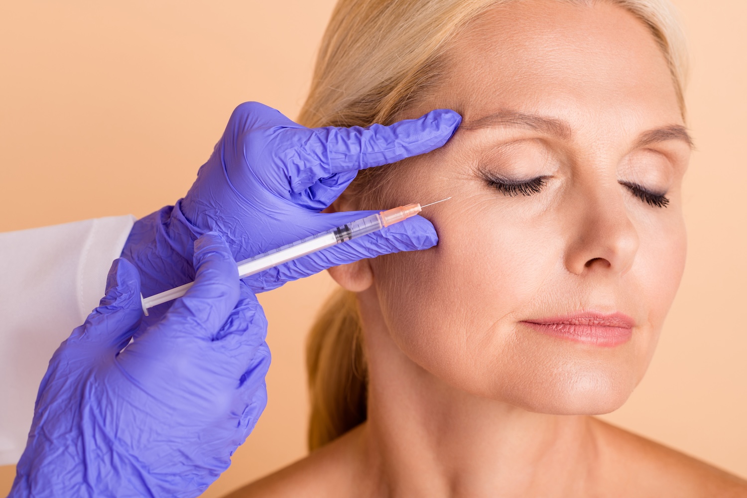 Gloved hands holding a syringe near the outer eye area of a woman with closed eyes against a neutral background.