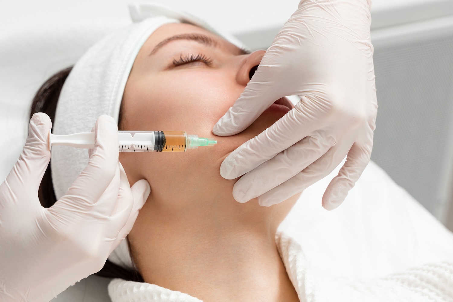 Gloved hands holding a syringe positioned at a woman’s cheek while she lies on a treatment bed with a headband.