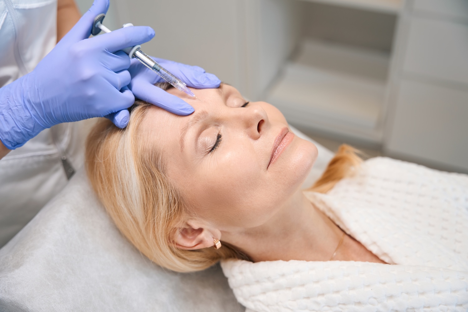 Gloved hands holding a syringe at a woman’s forehead as she lies on a treatment bed with eyes closed.