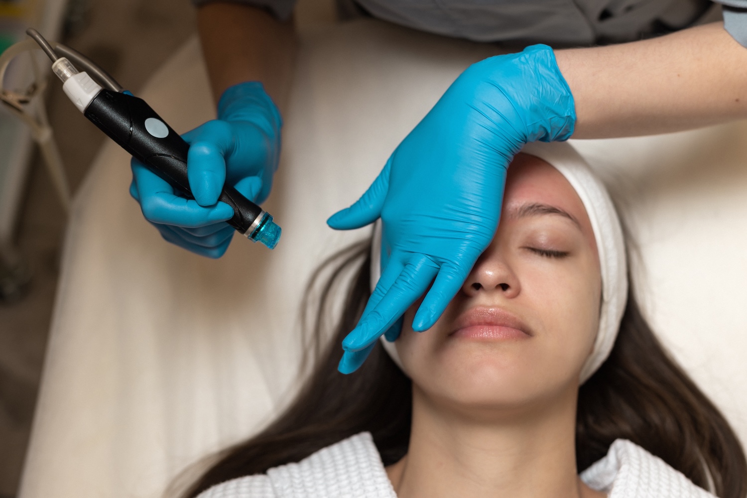 esthetician wearing blue gloves holding a hydrafacial device near a client’s forehead while the client lies with eyes closed