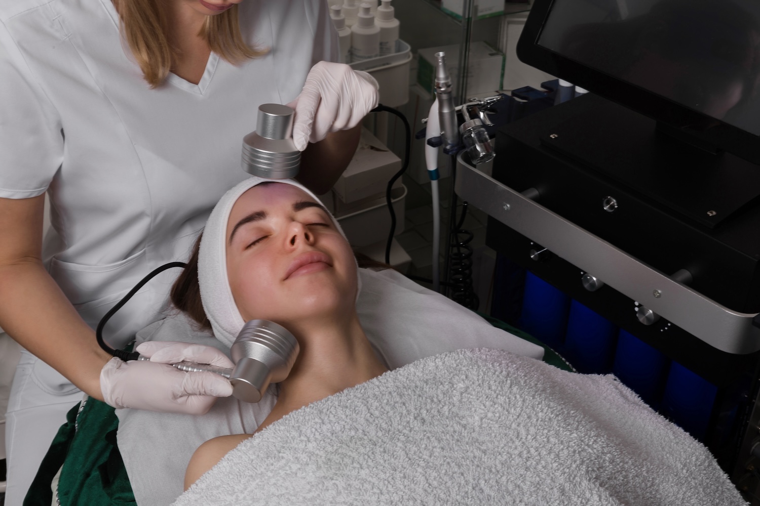 esthetician using two handheld devices on a client’s face and neck as the client lies on a treatment table with eyes closed