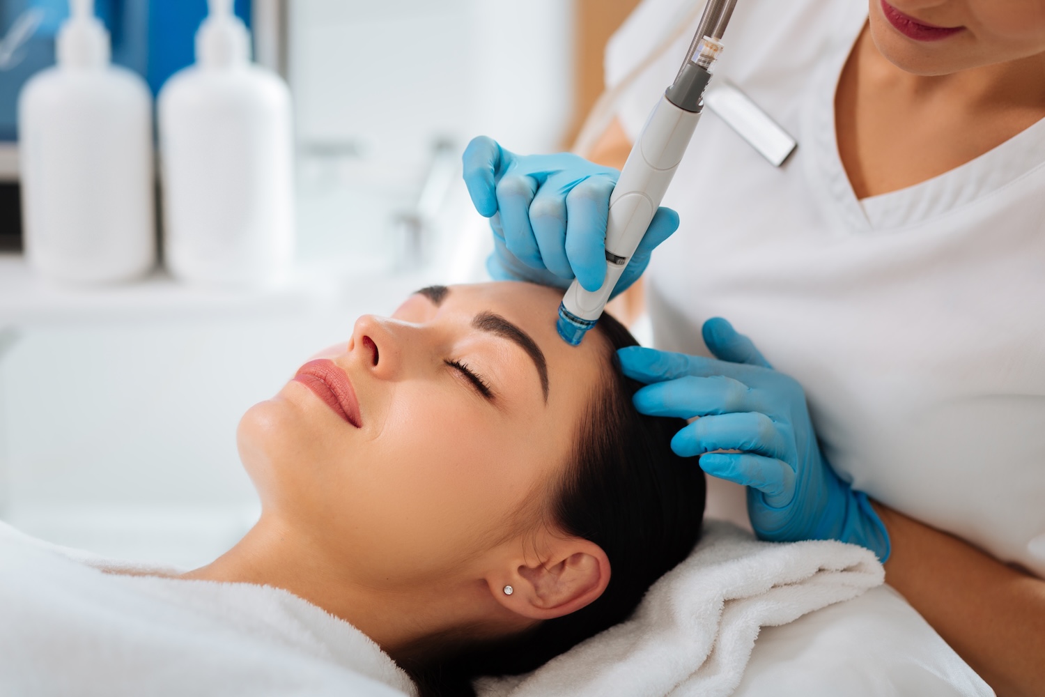 esthetician in blue gloves using a hydrafacial device on a client’s forehead as the client lies with eyes closed