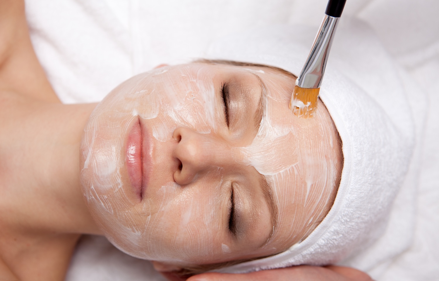 a woman receiving a facial as a creamy mask is applied with a brush while she relaxes with her eyes closed