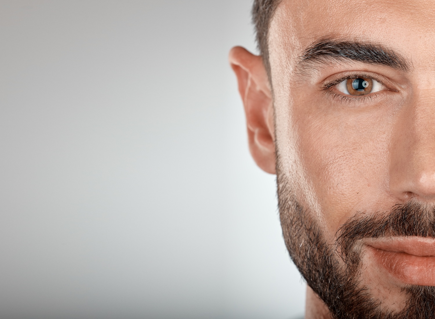close-up of the right side of a man's face showing his eye, eyebrow, cheek, and beard against a plain background