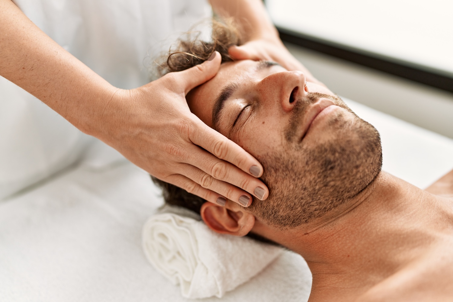 esthetician’s hands massaging the sides of a man’s face as he lies with eyes closed on a white towel