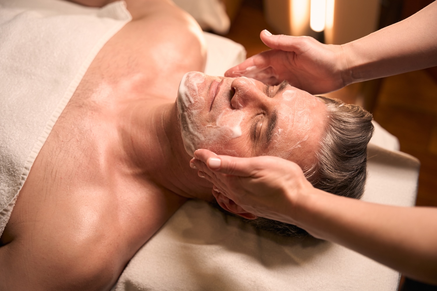 a man lying on a treatment table with his eyes closed while a practitioner applies a white facial cream to his face