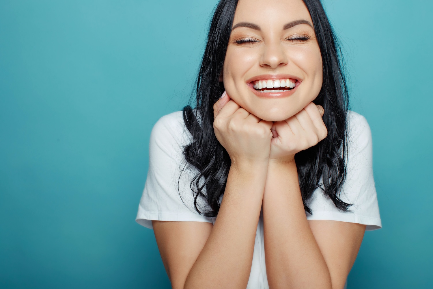 woman smiling with her eyes closed while resting her chin on her hands against a blue background