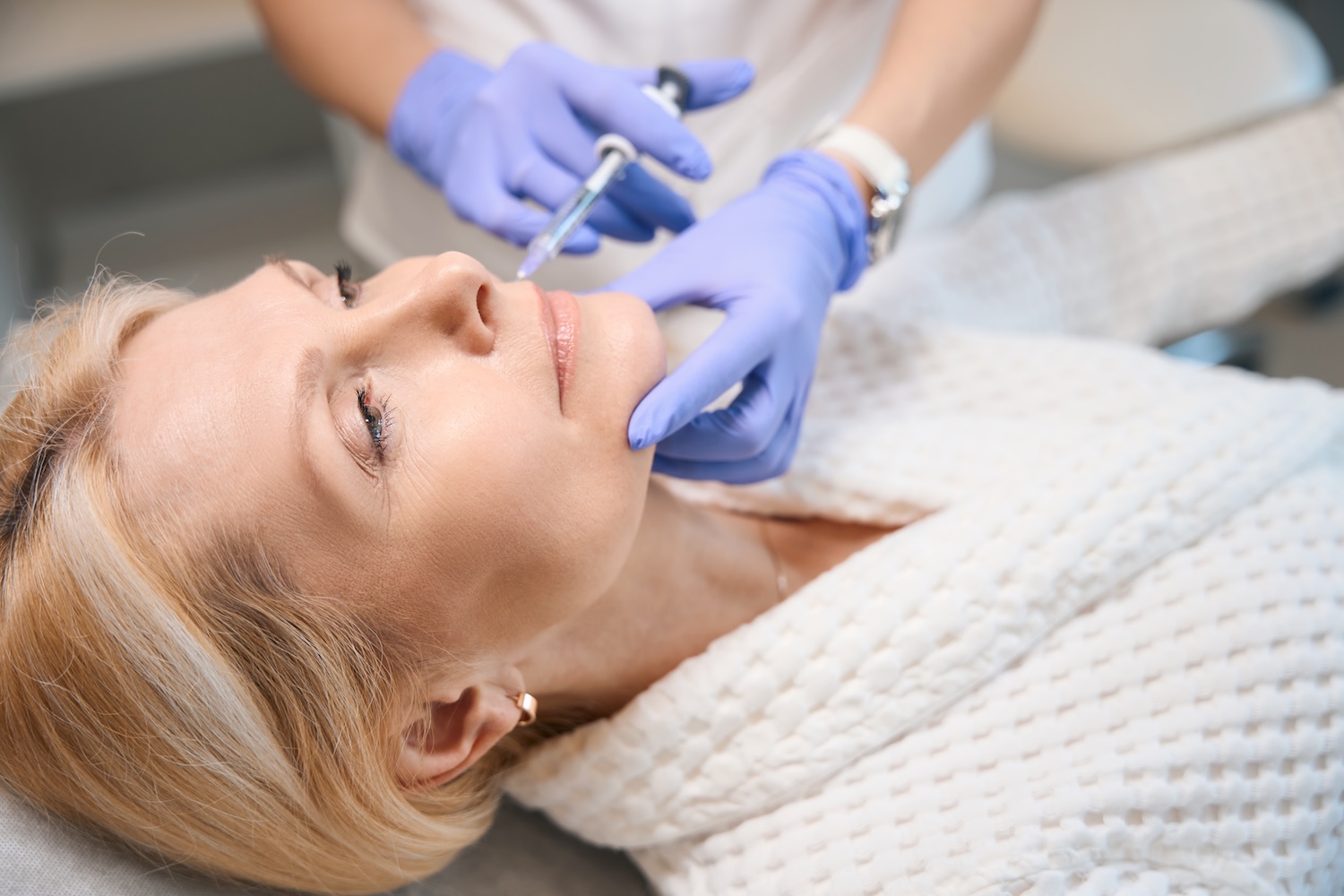 Gloved hands holding a syringe near a woman’s jawline as she lies on a treatment bed wearing a robe.