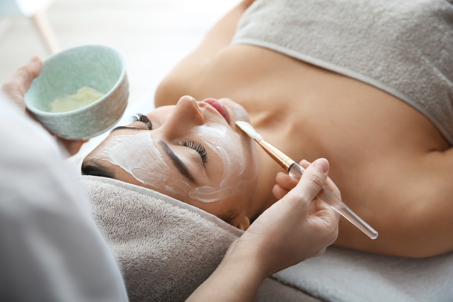 a practitioner uses a brush to apply a white facial mask to a woman's face as she lies on a treatment table