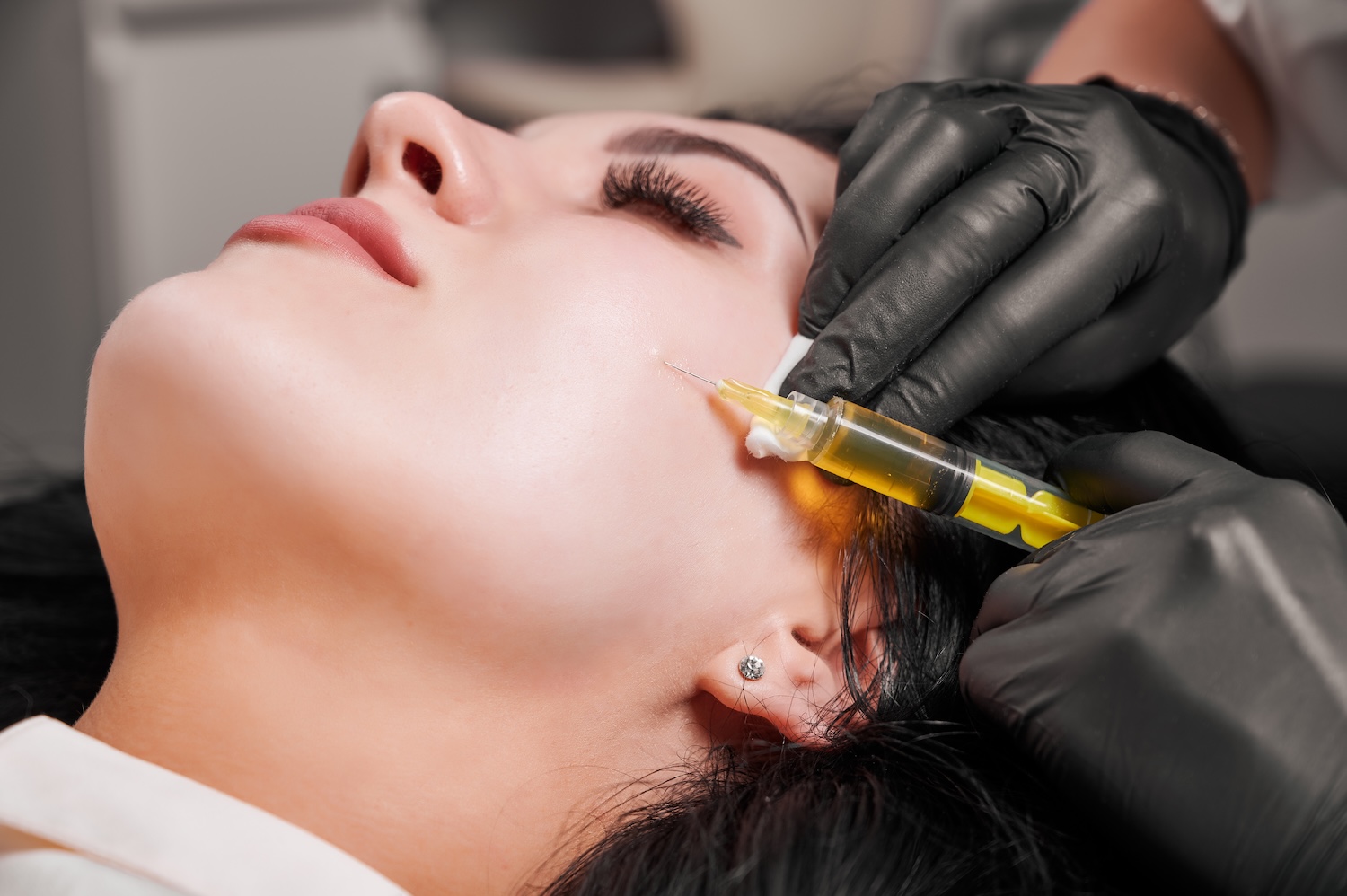 close-up of gloved hands holding a syringe with yellow fluid near a woman’s cheek as she lies back