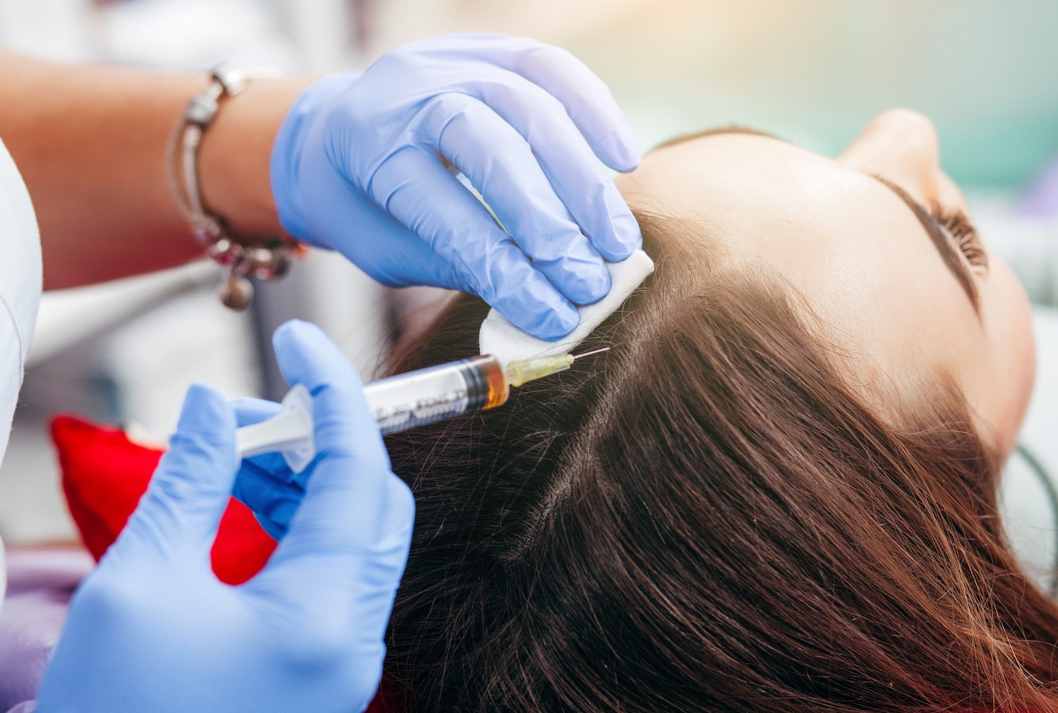 gloved hands parting a woman’s hair and holding a syringe near her scalp