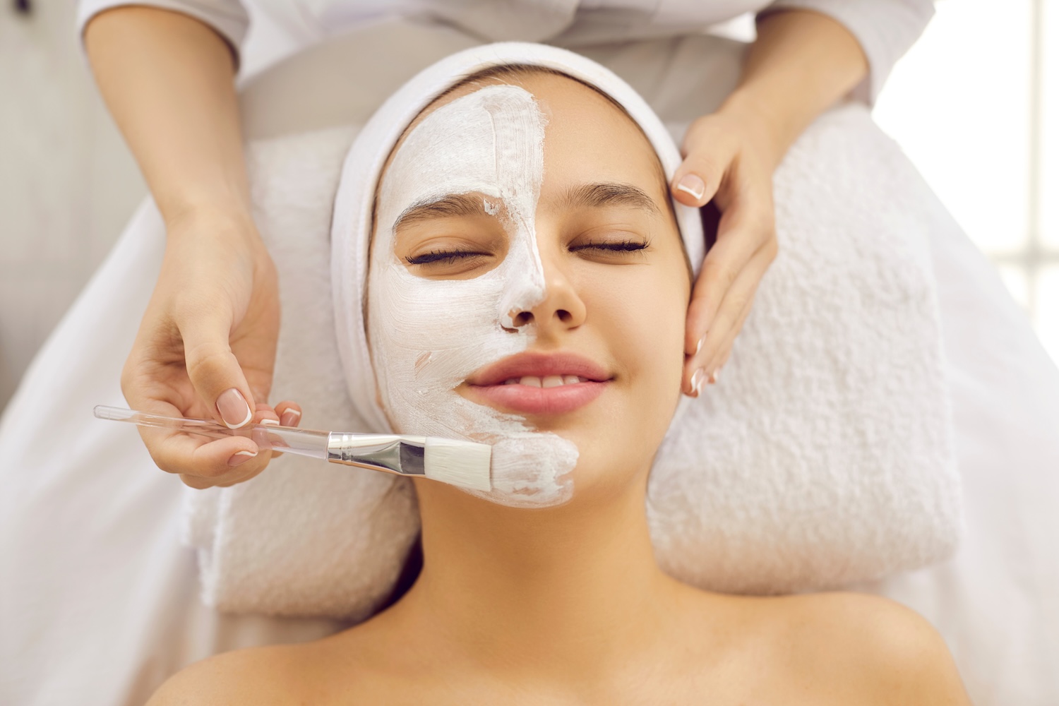 esthetician applying a white facial mask with a brush to a smiling teen girl during a relaxing skincare treatment