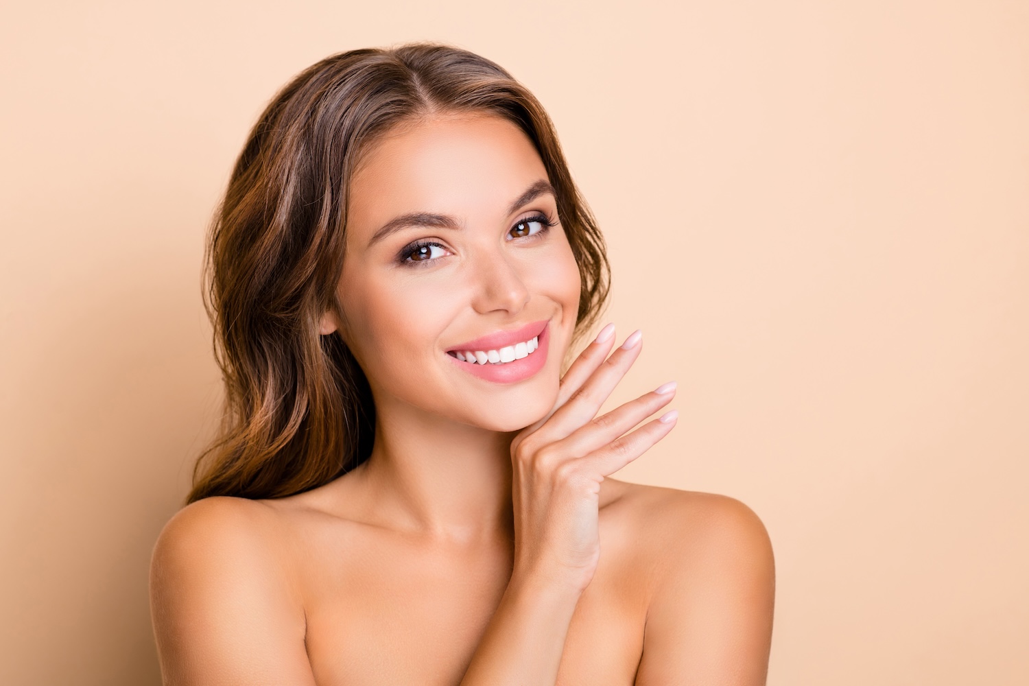 Woman smiling while resting her hand against her cheek, bare shoulders visible, against a solid peach background.
