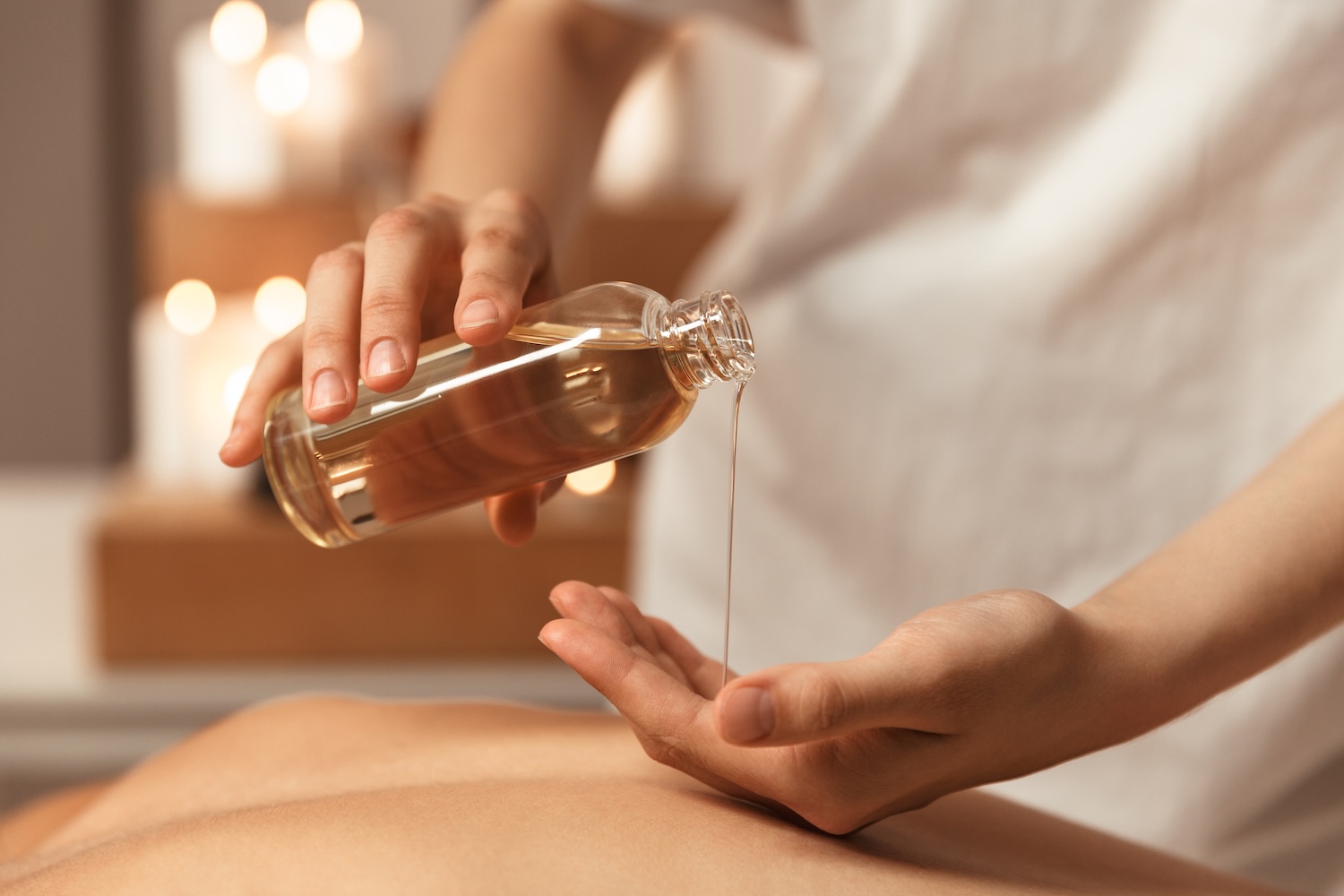therapist pouring massage oil from a glass bottle into their hand above a client’s back