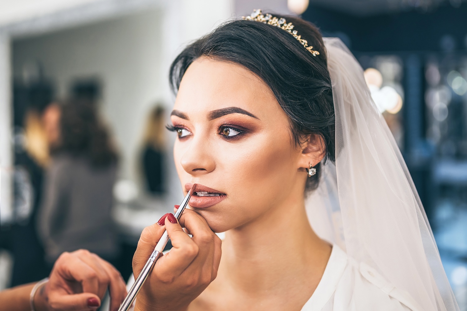 Makeup artist applying lipstick to a bride wearing a veil and headpiece, with completed eye makeup and hair styled up.
