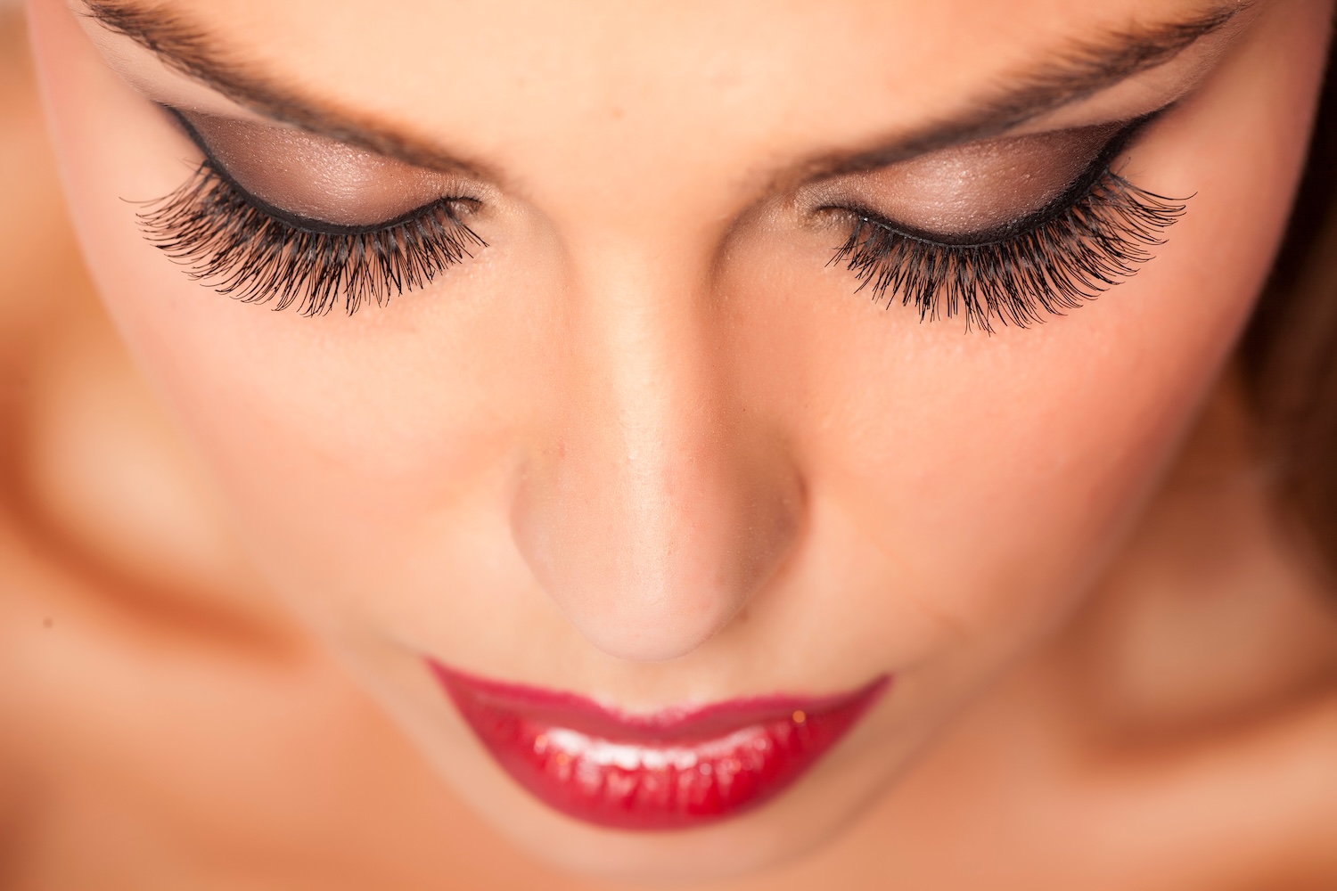 Top-down close-up of a person with closed eyes showing long, dark false eyelashes, eye makeup, and red lipstick.