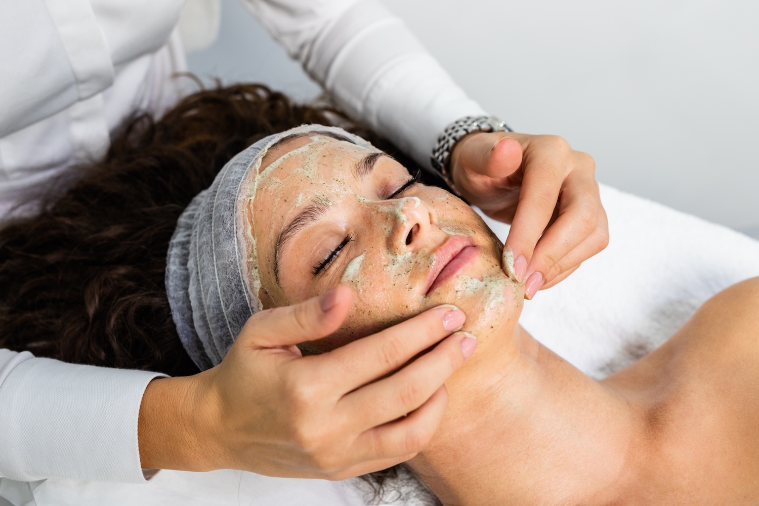 Practitioner applying a textured facial mask to a woman’s face as she lies on a treatment bed with eyes closed and a headband on.