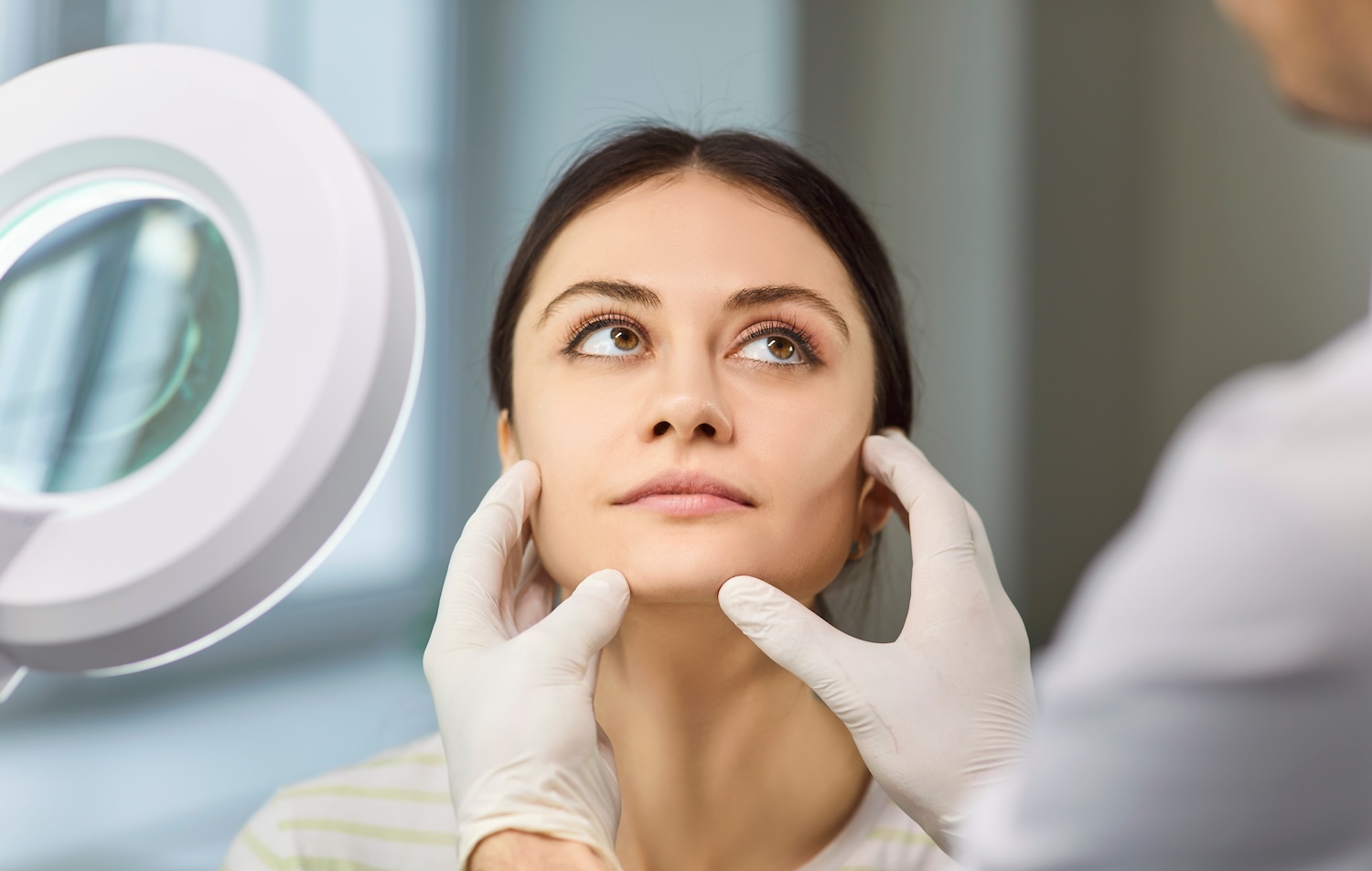 A woman looks upward while a practitioner wearing gloves gently touches her jawline and chin during a facial evaluation.