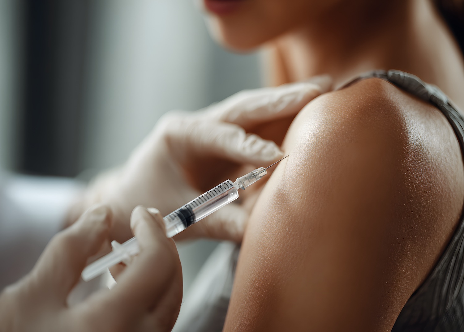 Gloved hands holding a syringe as a needle is positioned against a person’s shoulder for an injection.