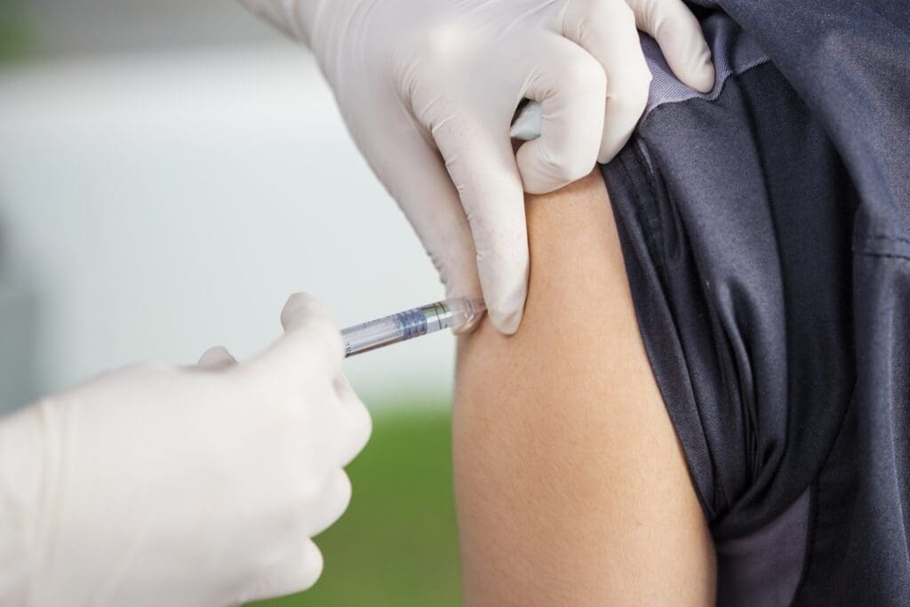 Gloved hands administer an injection into a person’s upper arm near the shoulder, with the syringe partially inserted into the skin.
