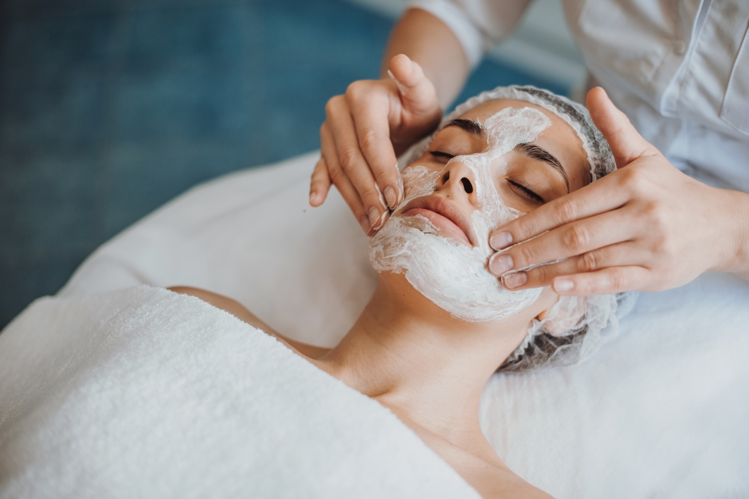 Practitioner applying a white facial mask with both hands to a woman’s face as she lies on a treatment bed with eyes closed.