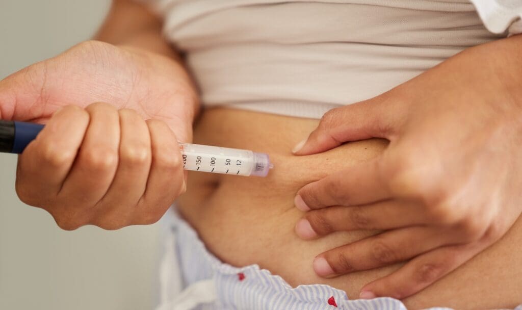 Hands holding an injection pen pressed against pinched skin on the lower abdomen.