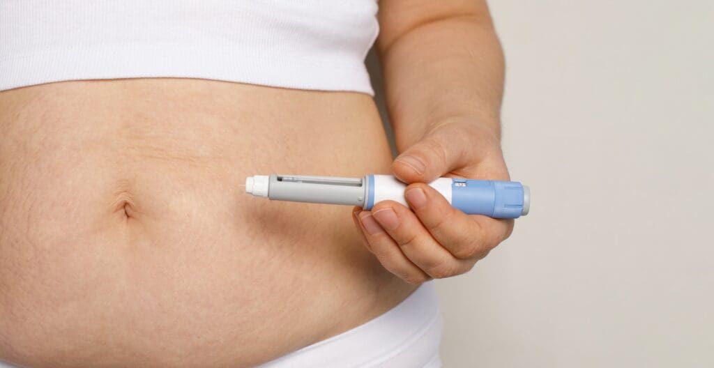 A person holds an injection pen against the skin of their lower abdomen near the navel.