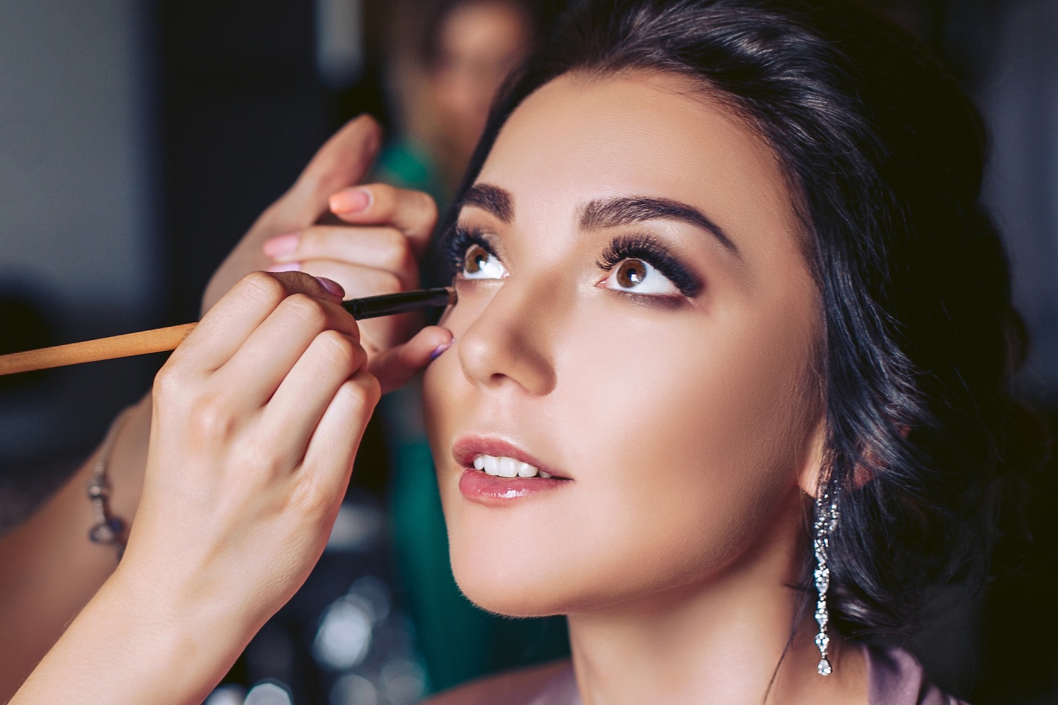 A makeup artist applies eyeliner beneath a woman’s eye using a brush while the woman looks upward.