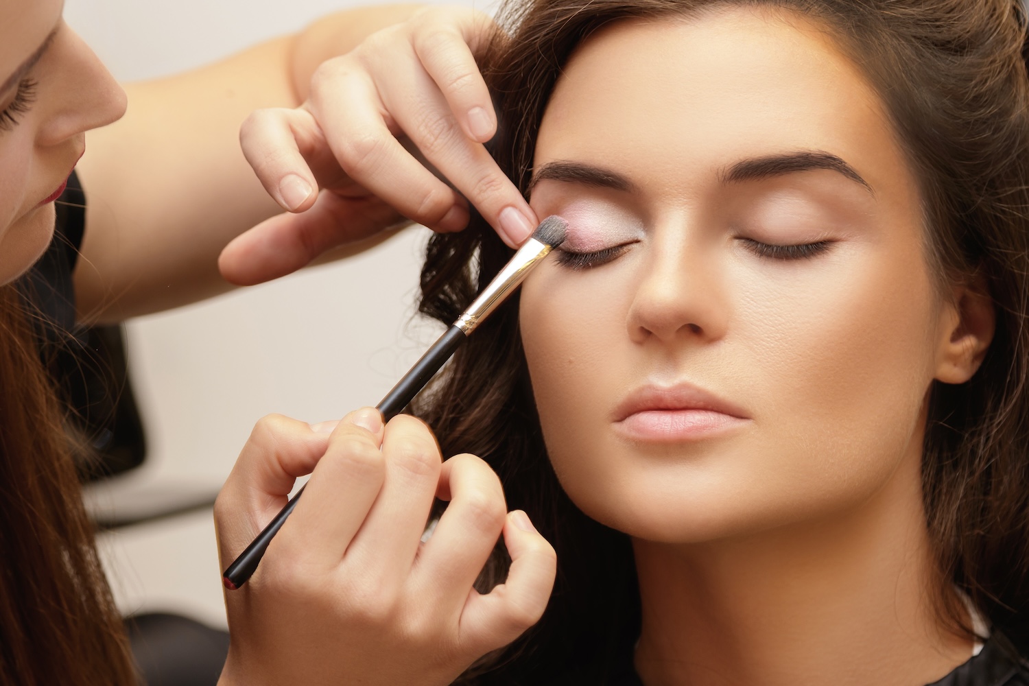 A makeup artist applies eyeshadow to a woman’s closed eyelid using a brush while gently lifting the skin above the eye.