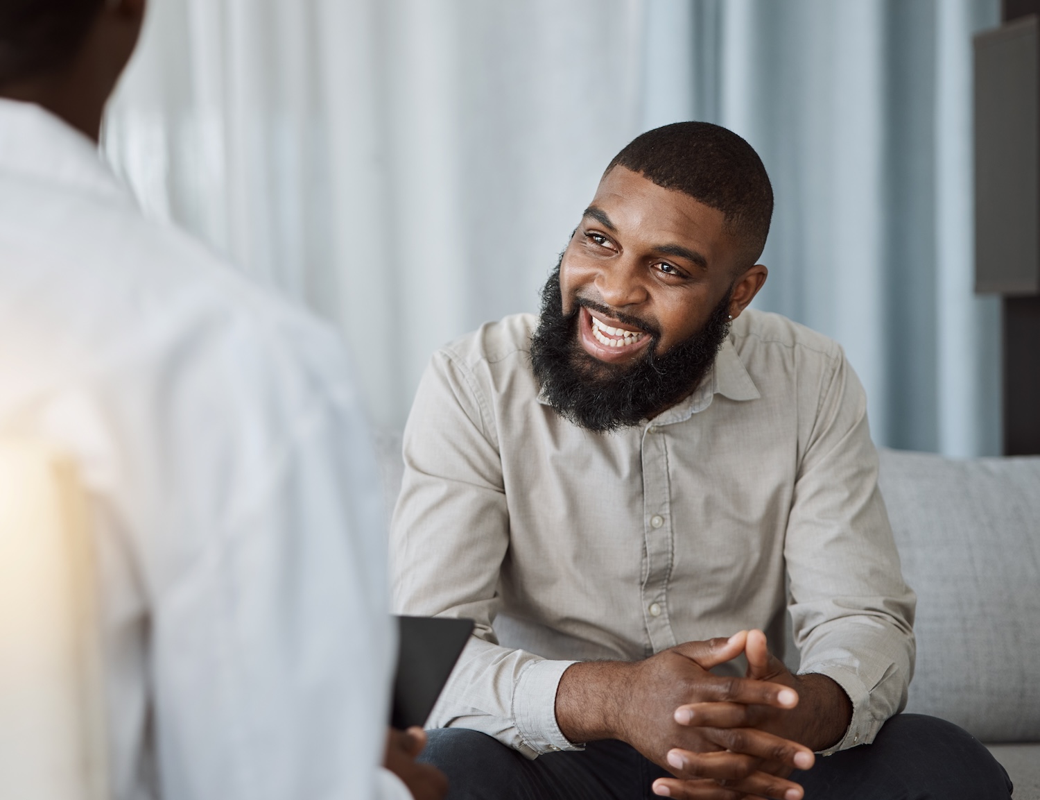 A bearded man sits with hands clasped, speaking to a clinician seated opposite during an in-office consultation.