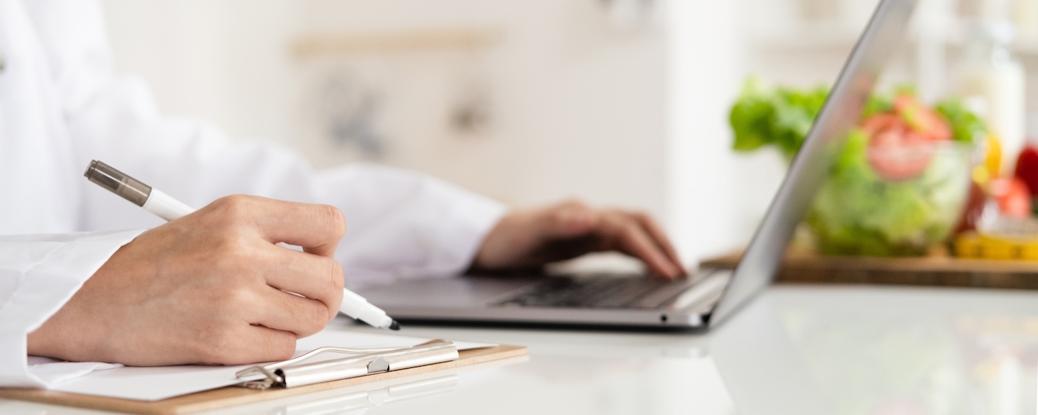 Healthcare professional writing on a clipboard while working on a laptop, with fresh vegetables blurred in the background.