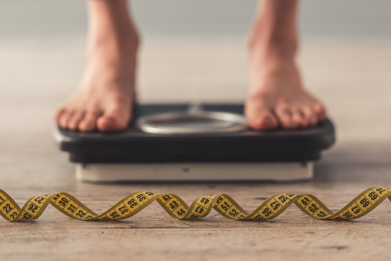 Bare feet standing on a bathroom scale, with a yellow measuring tape laid out on the floor in front.