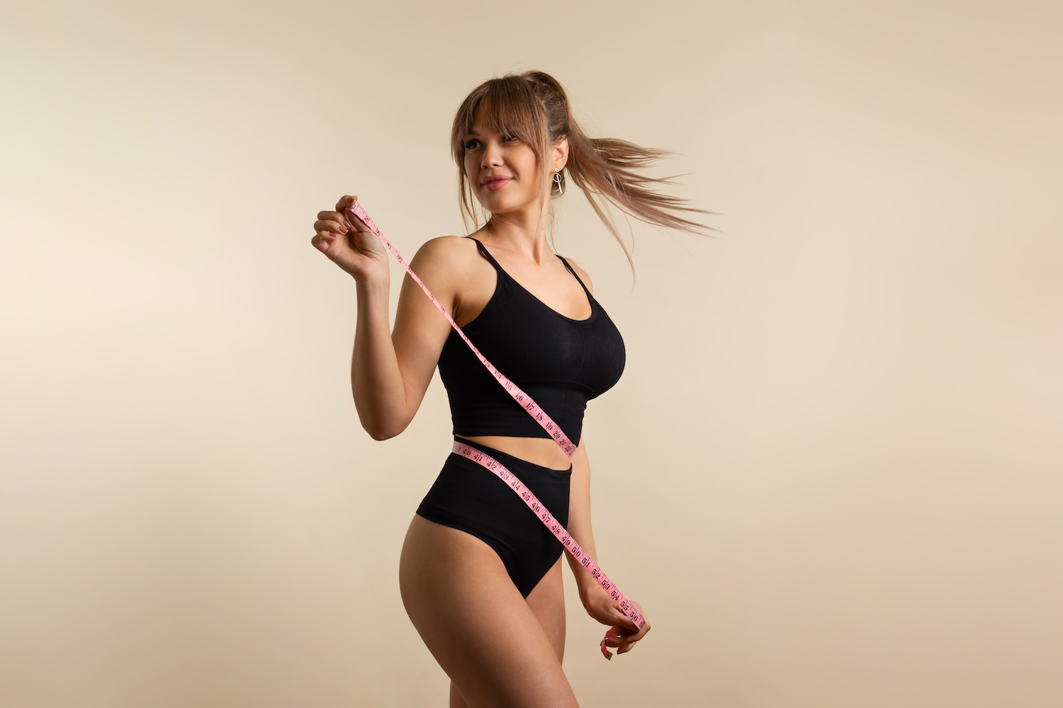 Woman wearing black athletic clothing holds a pink measuring tape around her waist while standing against a plain background