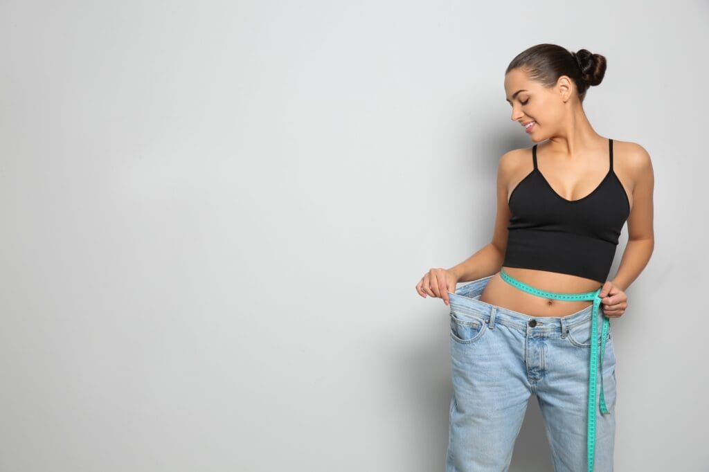 Woman in a black crop top holding out the waistband of oversized jeans with a tape measure around her waist.