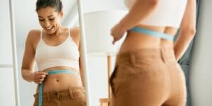 Woman standing in front of a mirror measuring her waist with a blue tape measure.